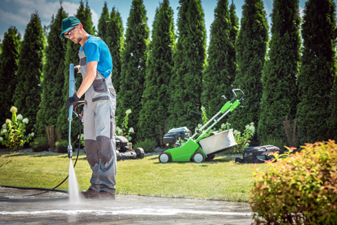 man using a pressure washer on pavement in a garden surrounded by tall shrubs and lawn care equipment for outdoor maintenance and cleaning 2 solutions