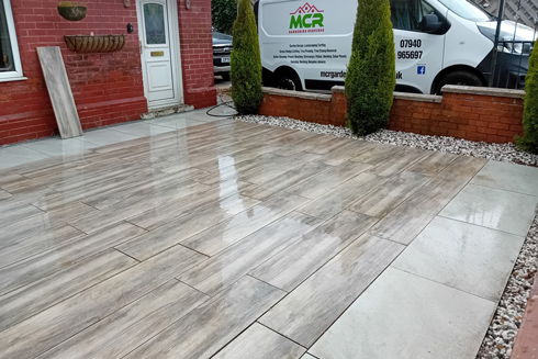 newly installed outdoor patio with wooden effect tiles and decorative stones featuring green shrubs and a delivery van in the background suitable for three season enjoyment
